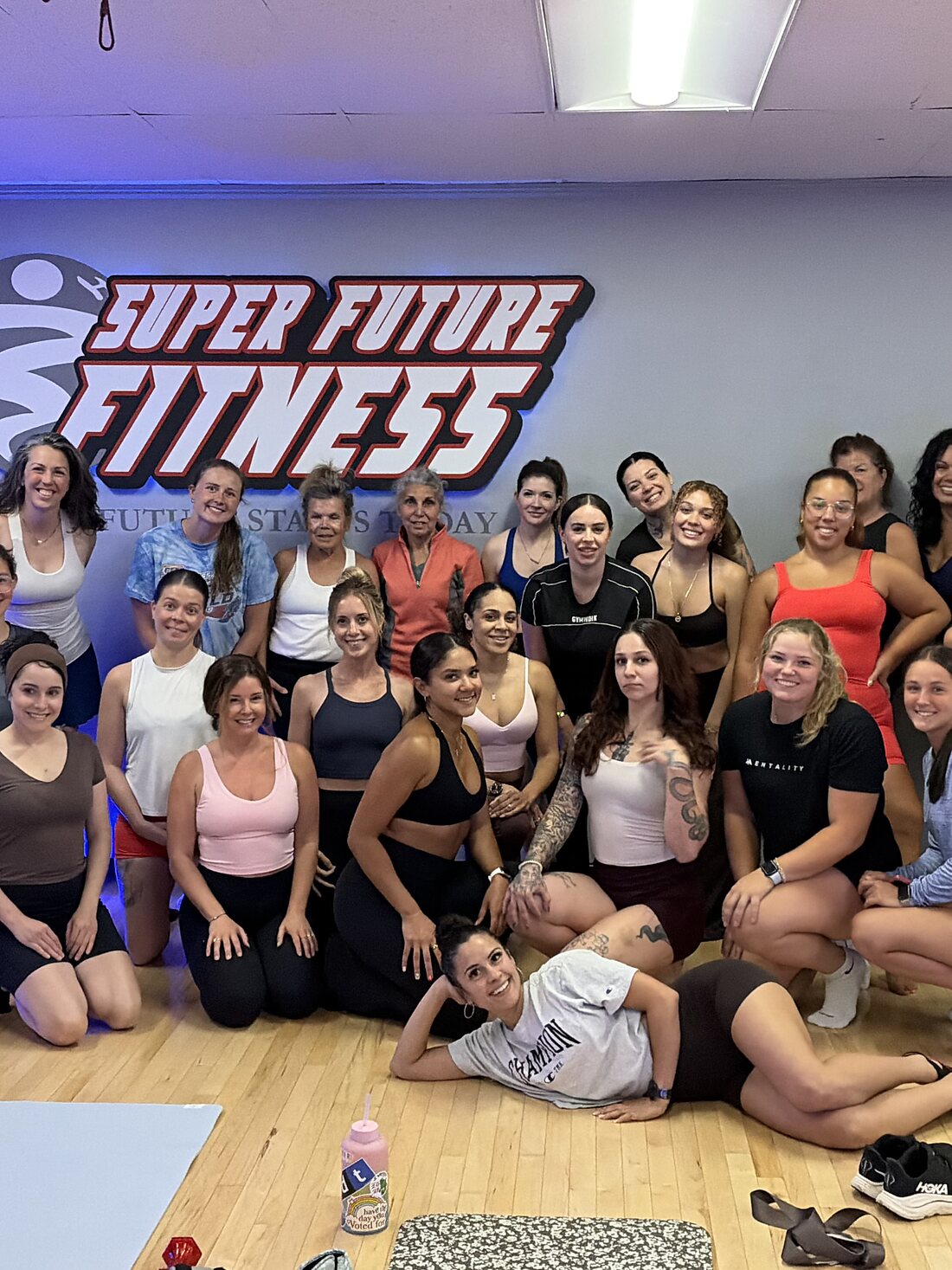 A full class of members smiling together under the lit Super Future Fitness logo right after their workout